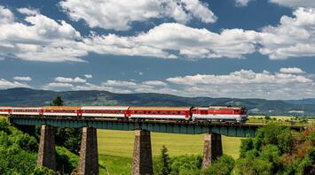 A red passenger train crossing a tall viaduct in a lush, green landscape under a cloudy sky