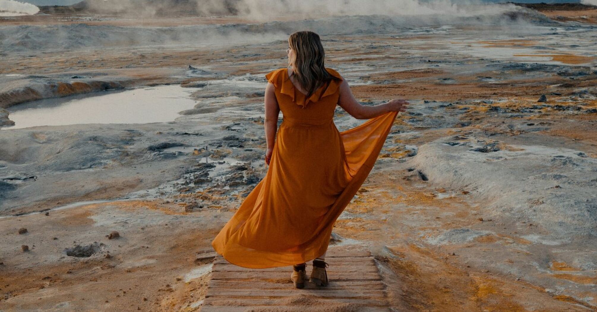 Woman in a flowing yellow dress walking along a geothermal landscape with steam rising in the background