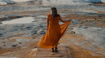 Woman in a flowing yellow dress walking along a geothermal landscape with steam rising in the background