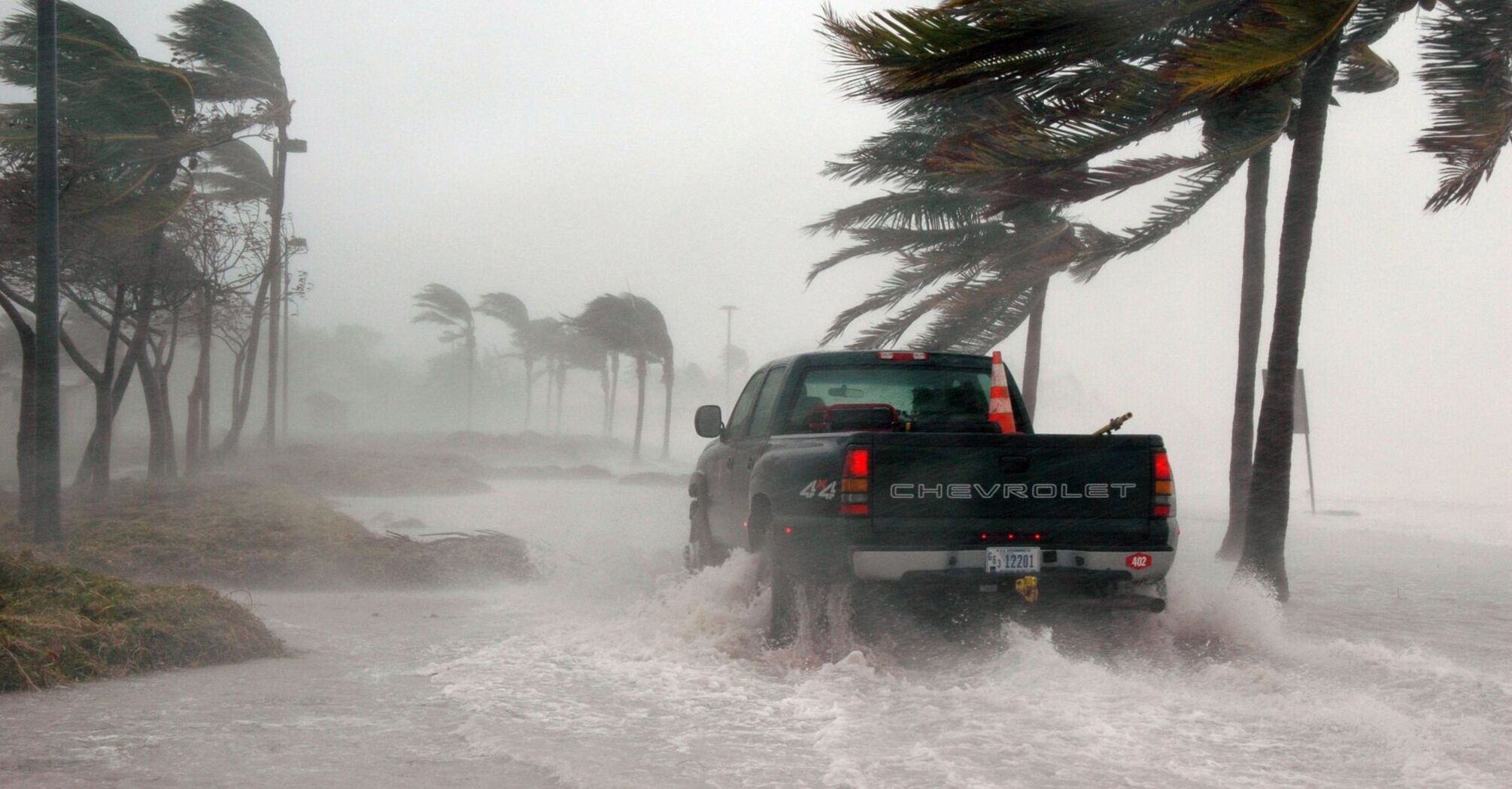 Truck driving through flooded road during a hurricane with strong winds bending palm trees