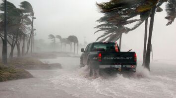 Truck driving through flooded road during a hurricane with strong winds bending palm trees
