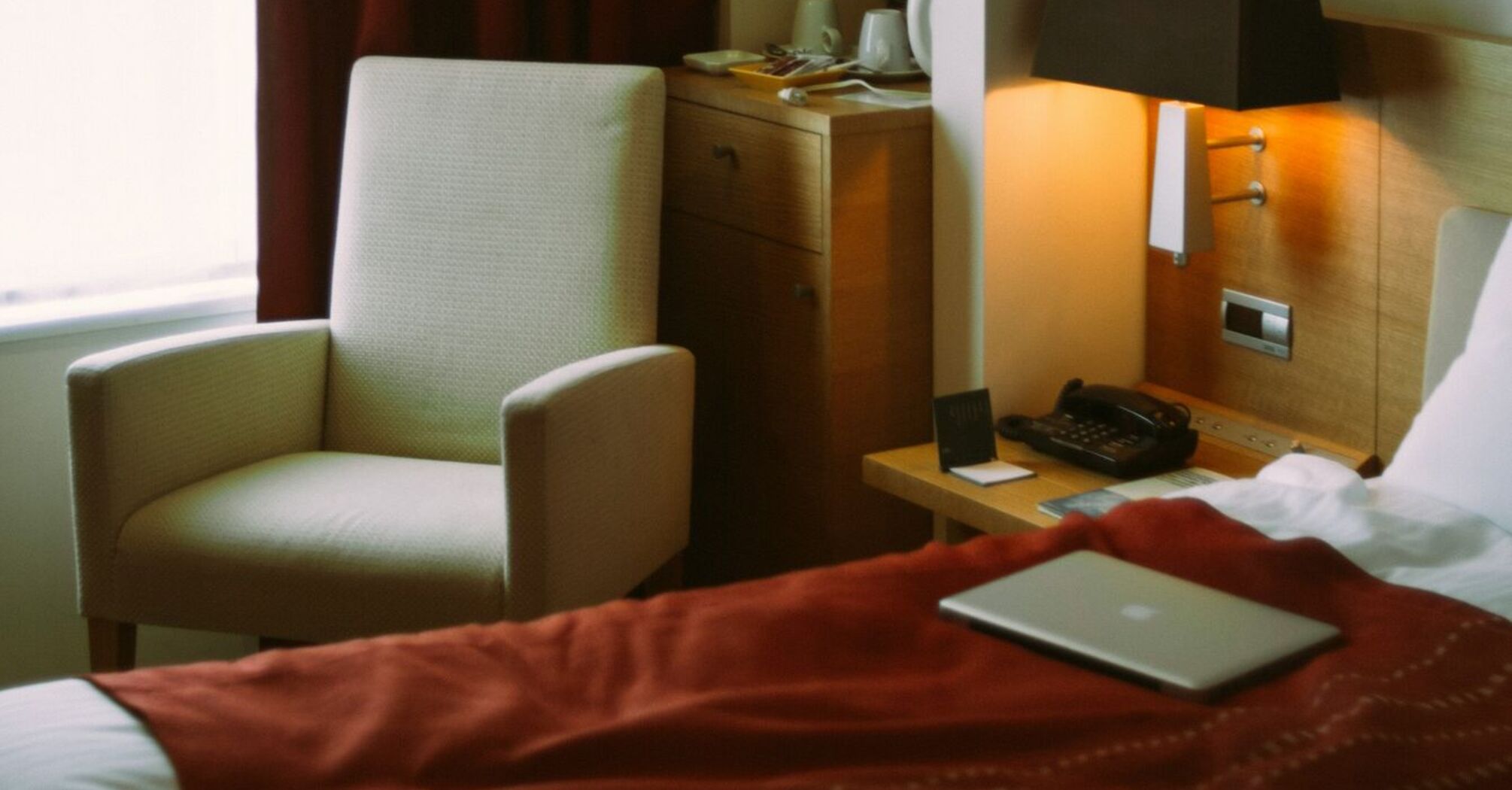 A cozy hotel room with a bed, a white armchair, and a desk lamp next to a window with red curtains. A laptop sits on the bed, with soft lighting adding to the warm ambiance