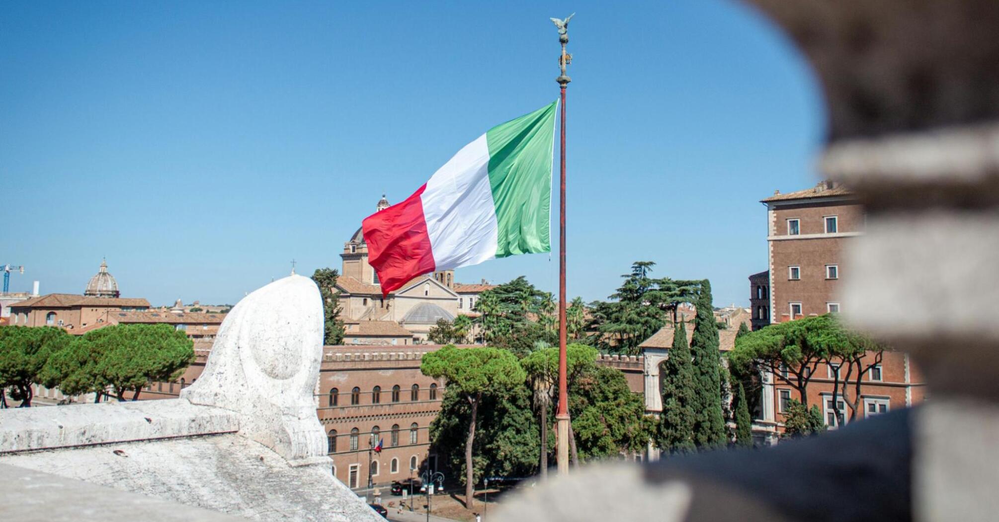 The Italian flag waving on a flagpole against a clear blue sky, with historic buildings and trees in the background