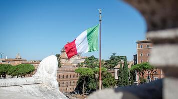 The Italian flag waving on a flagpole against a clear blue sky, with historic buildings and trees in the background