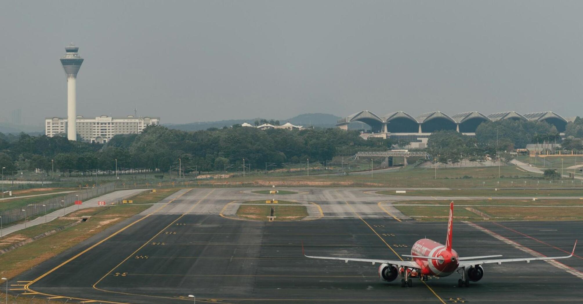 A red AirAsia airplane taxiing on the runway at an airport with a control tower and terminal buildings in the background