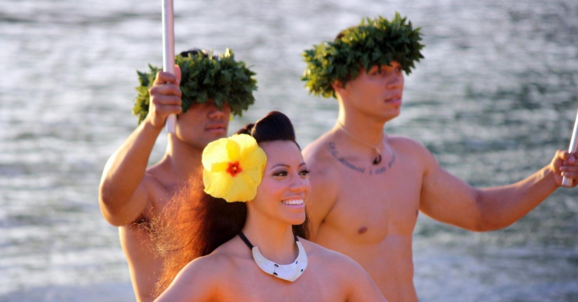 Performers in traditional Polynesian attire on a canoe, one woman smiling brightly, surrounded by two men with ceremonial headgear