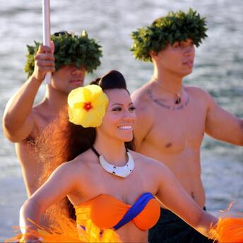 Performers in traditional Polynesian attire on a canoe, one woman smiling brightly, surrounded by two men with ceremonial headgear