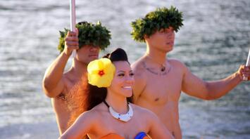 Performers in traditional Polynesian attire on a canoe, one woman smiling brightly, surrounded by two men with ceremonial headgear