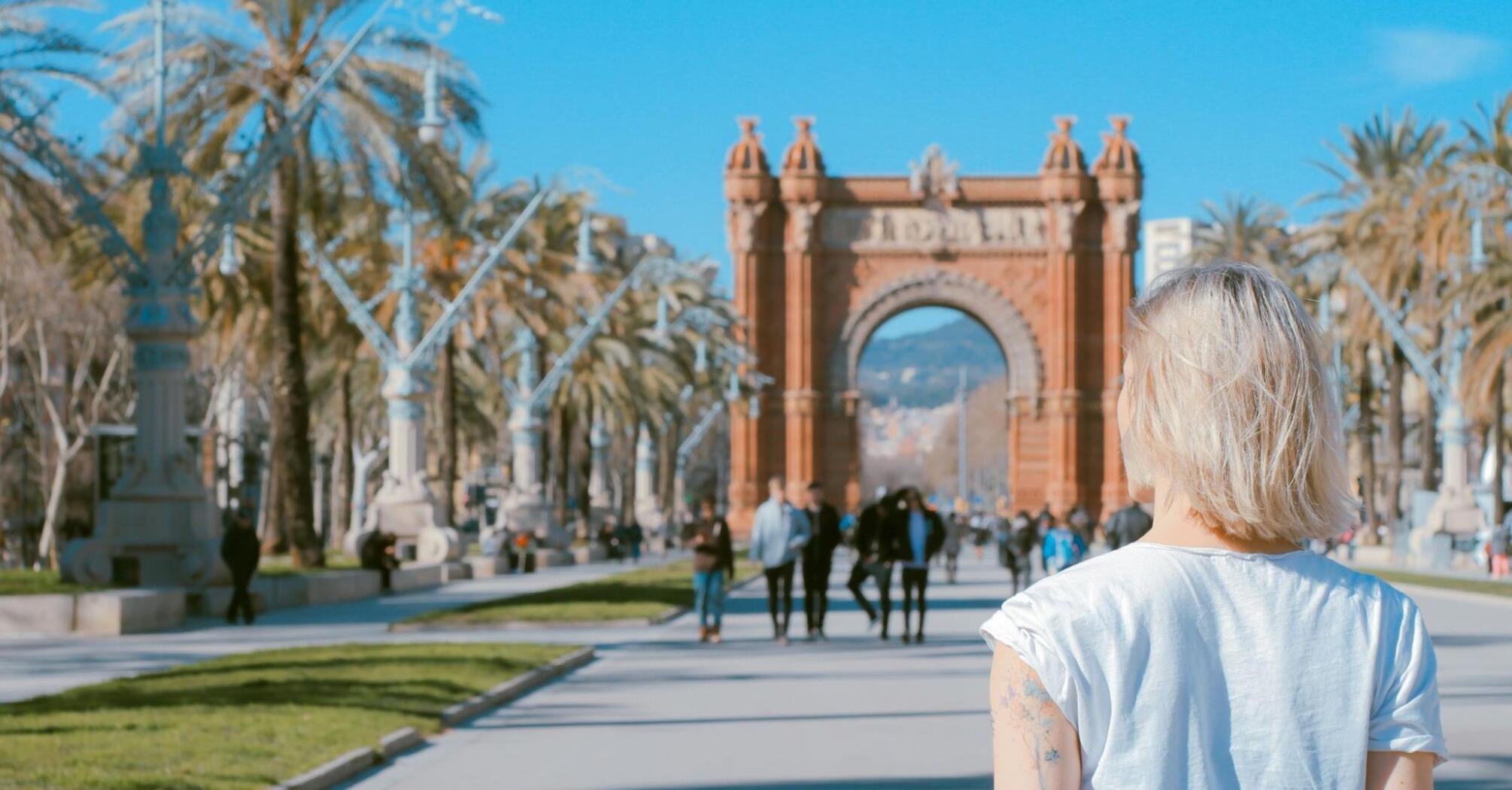 A woman with short blonde hair walks towards the Arc de Triomf in Barcelona, Spain, surrounded by palm trees on a sunny day