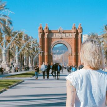 A woman with short blonde hair walks towards the Arc de Triomf in Barcelona, Spain, surrounded by palm trees on a sunny day