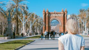 A woman with short blonde hair walks towards the Arc de Triomf in Barcelona, Spain, surrounded by palm trees on a sunny day