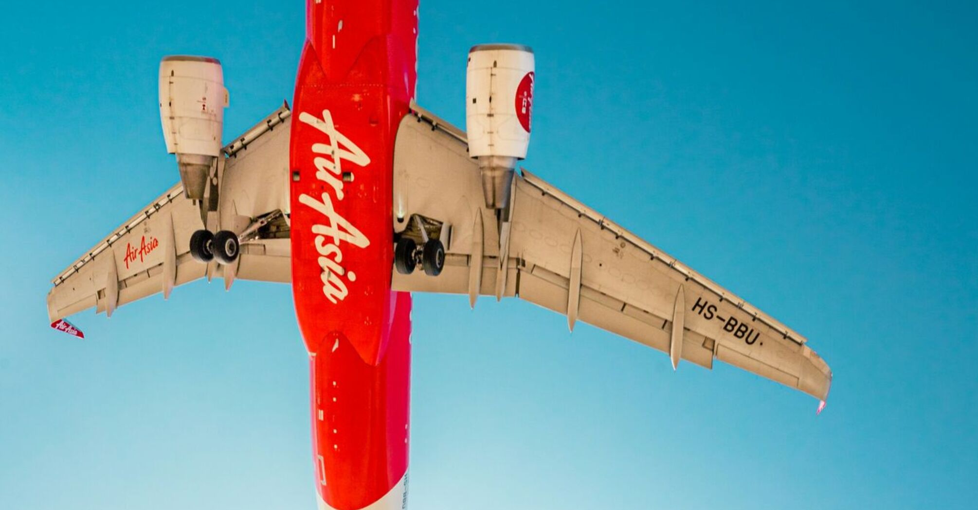 A woman in a floral dress stands beneath an AirAsia airplane as it flies low overhead, captured against a clear blue sky