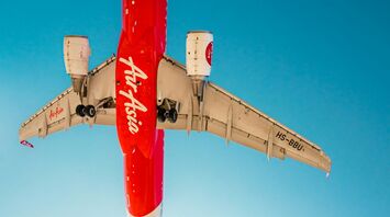 A woman in a floral dress stands beneath an AirAsia airplane as it flies low overhead, captured against a clear blue sky