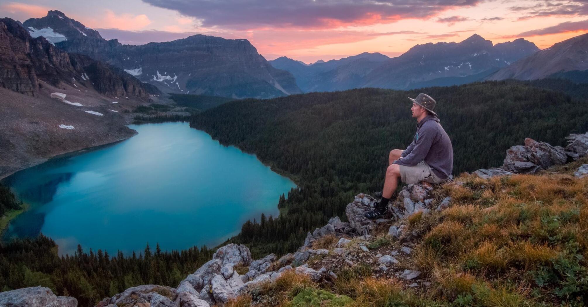 A hiker sits on a rocky outcrop overlooking a serene mountain lake surrounded by forest and mountains at sunset