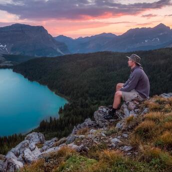 A hiker sits on a rocky outcrop overlooking a serene mountain lake surrounded by forest and mountains at sunset