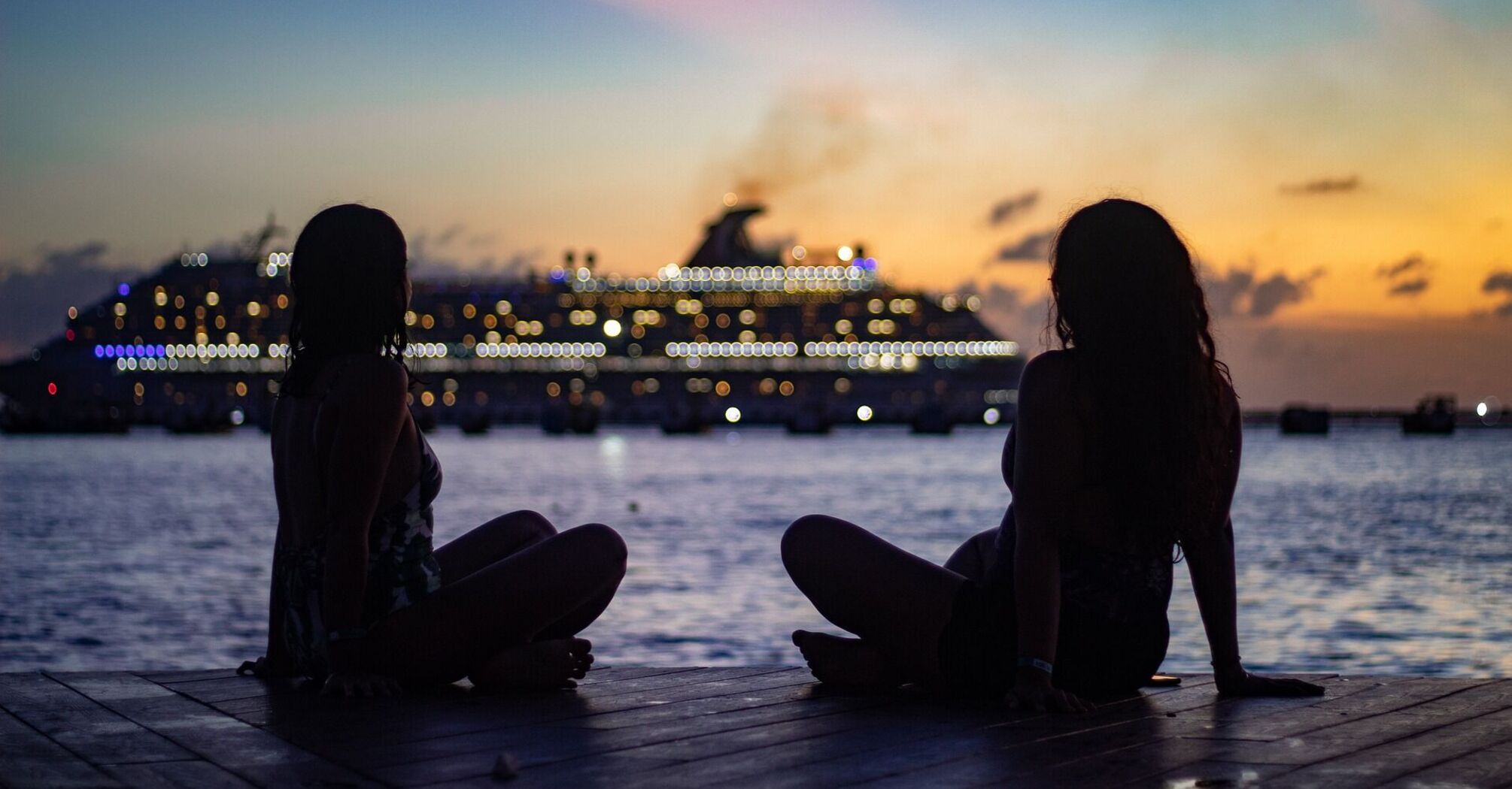 Two women sitting on a pier at sunset with a cruise ship in the background