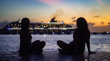 Two women sitting on a pier at sunset with a cruise ship in the background