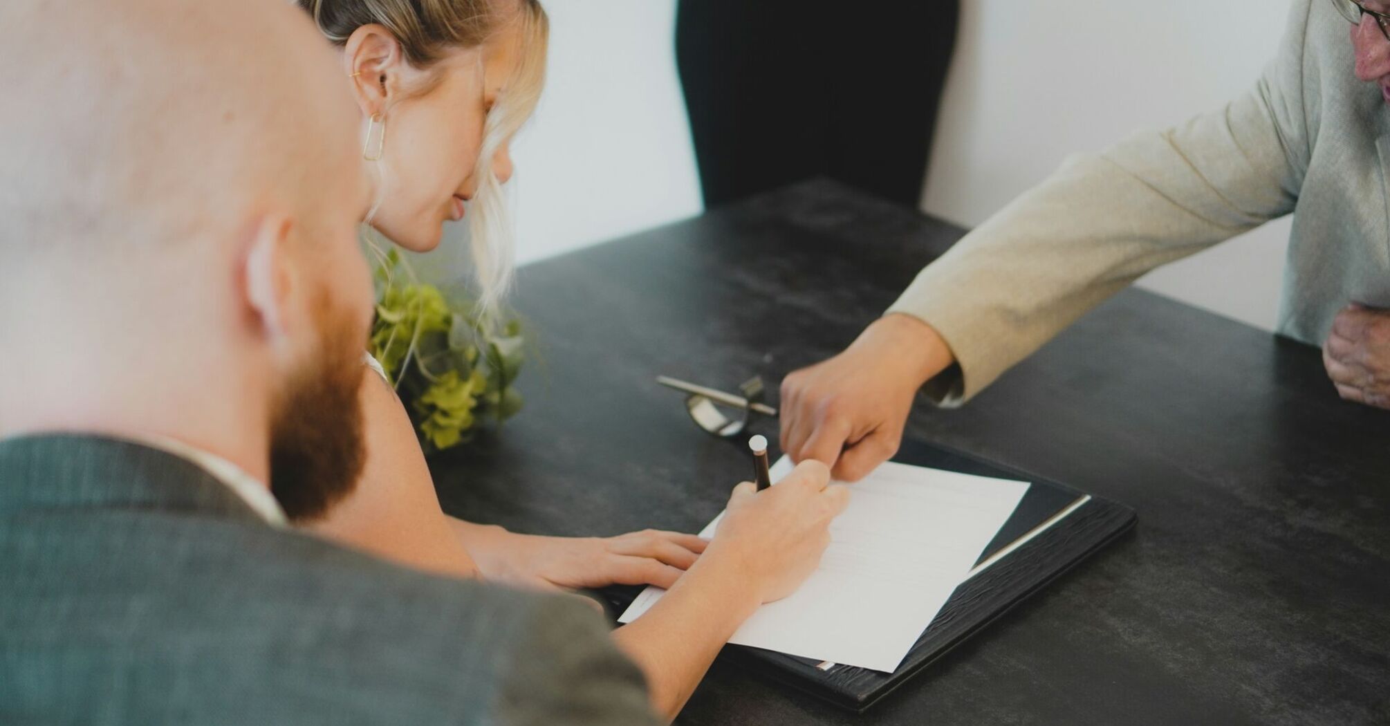 A woman signs a document during a formal meeting, with another person guiding her hand