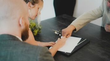 A woman signs a document during a formal meeting, with another person guiding her hand