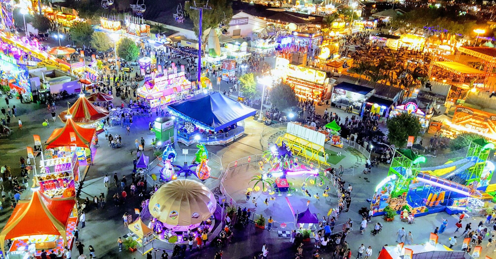 A vibrant nighttime view of a fair showcasing colorful rides, food stalls, and visitors enjoying the event