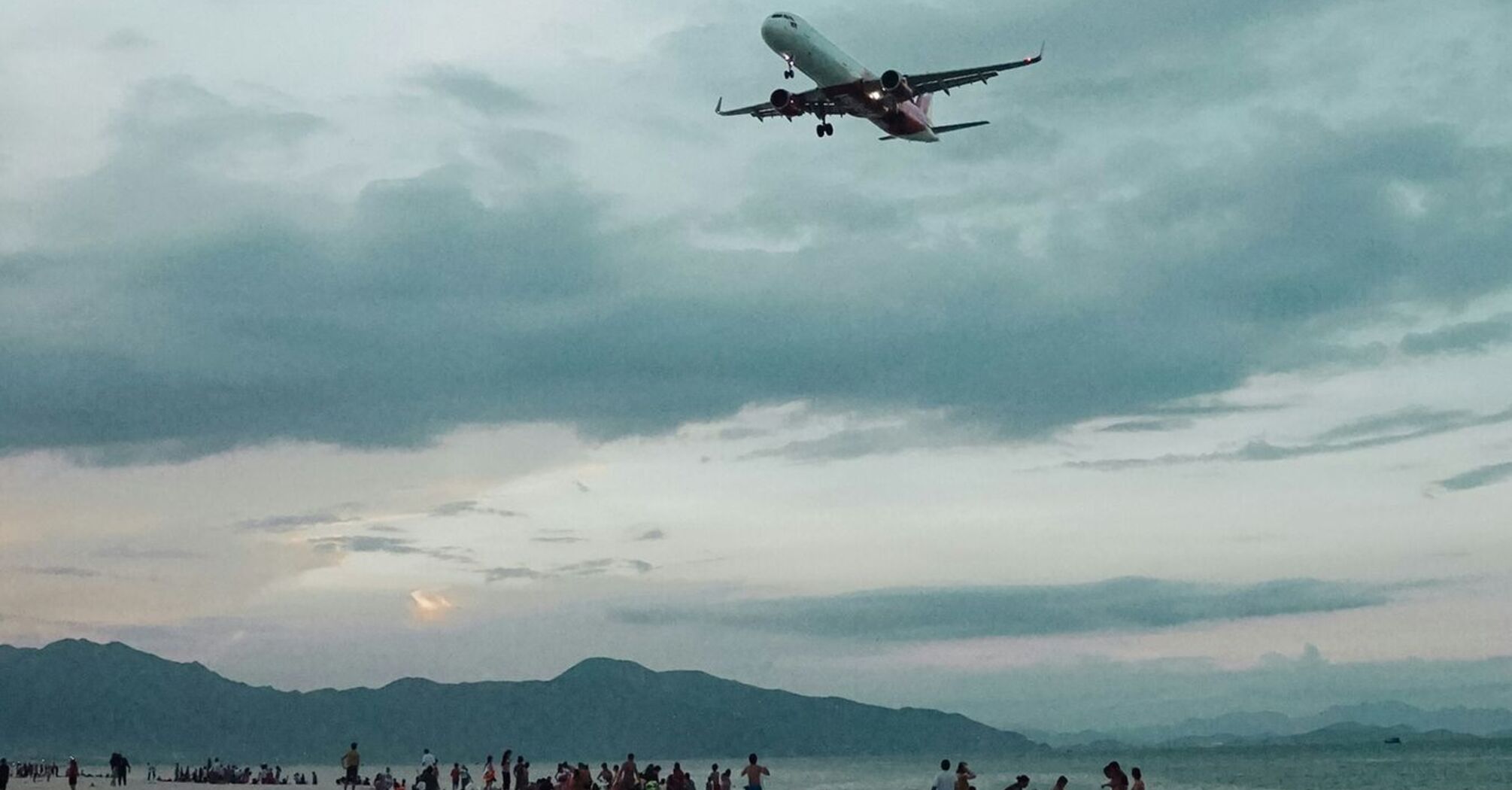 A Vietjet airplane flies over a beach, with people gathered on the sand and mountains in the distance under a cloudy sky