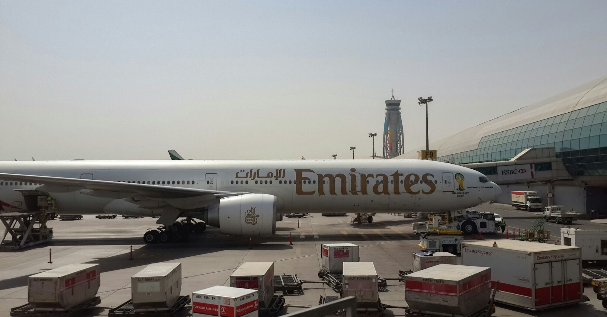 Emirates airplane stationed at the airport terminal, with cargo containers and airport equipment visible in the foreground