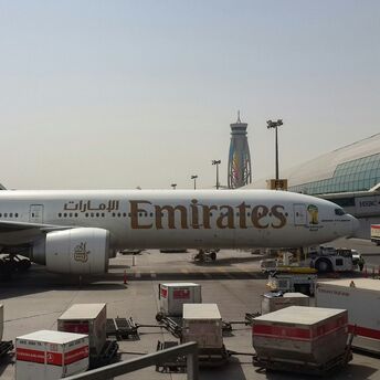 Emirates airplane stationed at the airport terminal, with cargo containers and airport equipment visible in the foreground