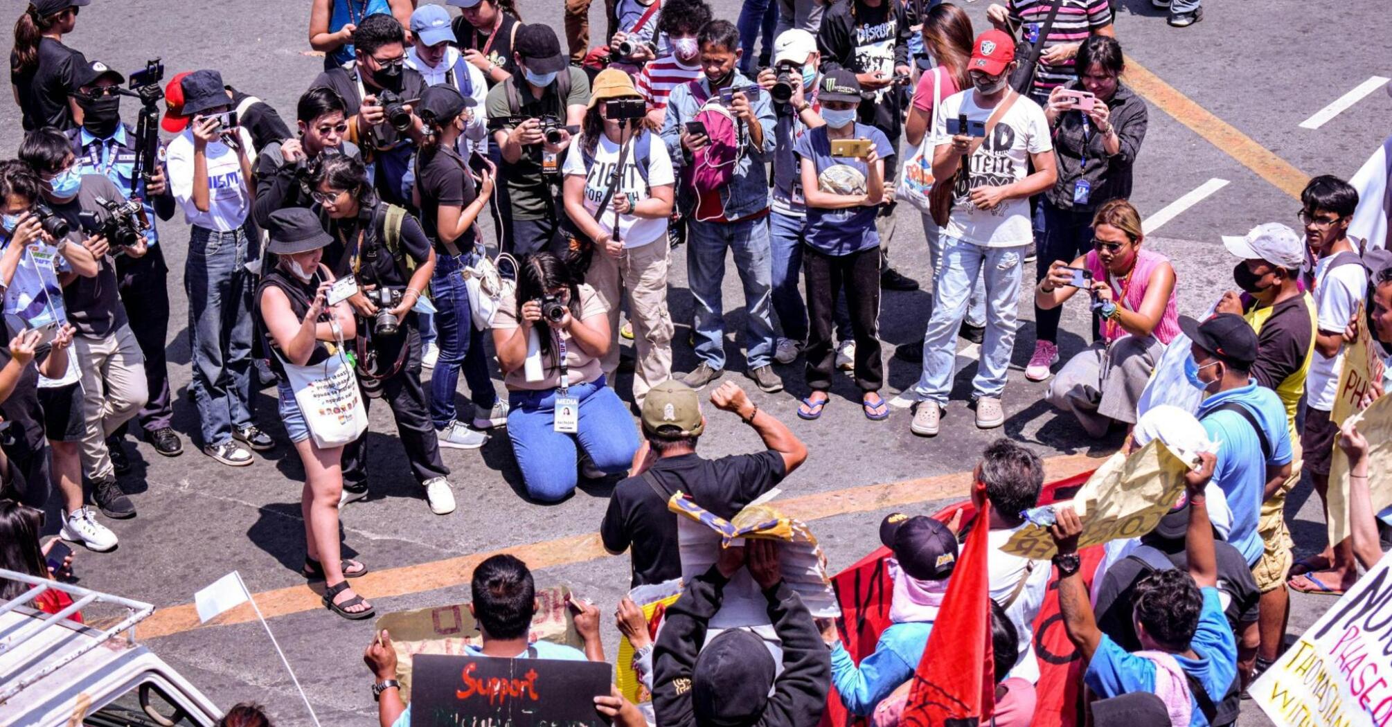 A large group of people, including photographers and protestors, gather in a public space during a demonstration, with some individuals holding signs and banners