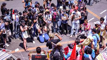A large group of people, including photographers and protestors, gather in a public space during a demonstration, with some individuals holding signs and banners