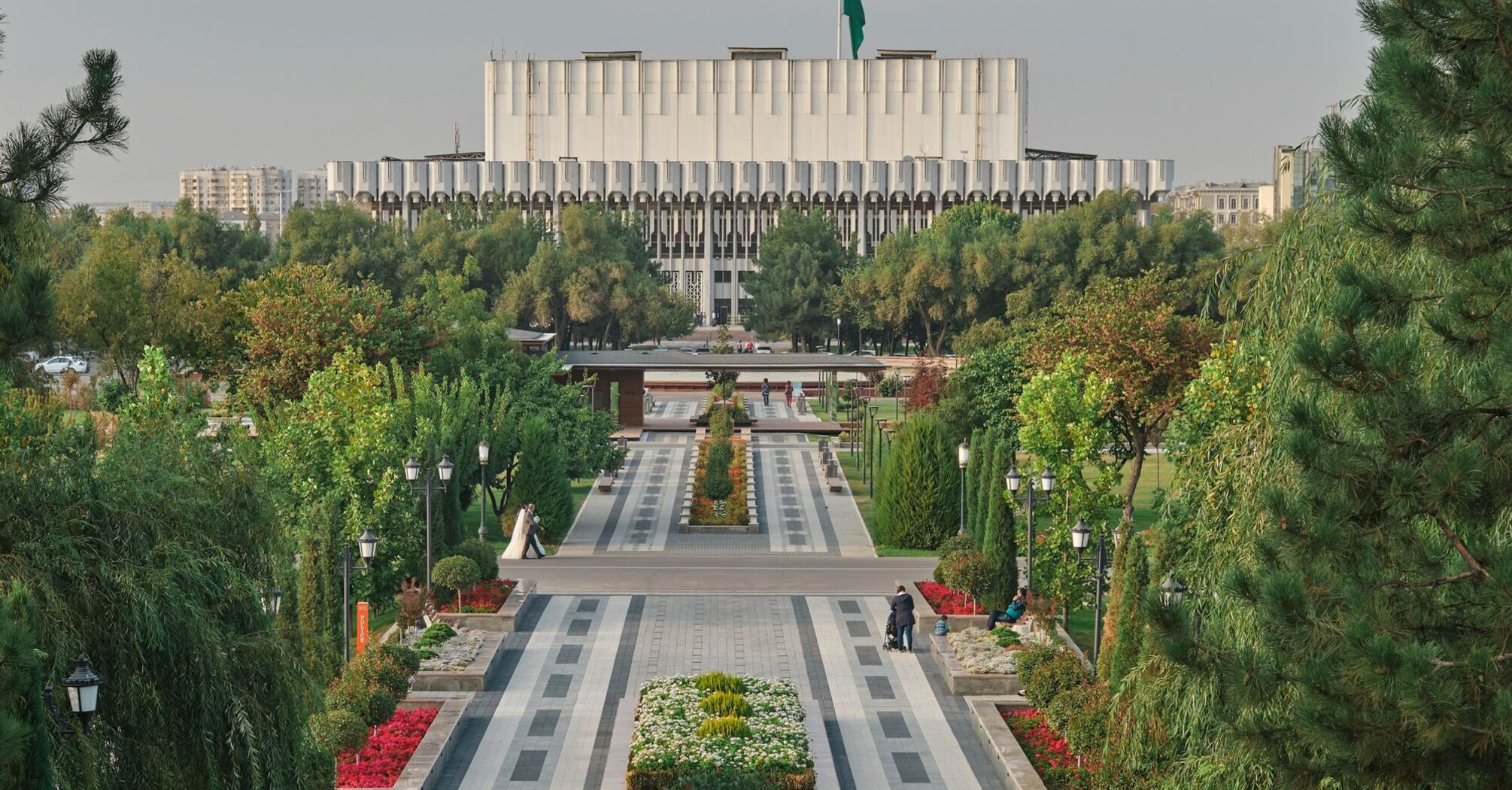 A beautifully landscaped park in Tashkent, Uzbekistan, with neatly arranged flower beds, paved pathways, and the Uzbekistan State Museum of History in the background