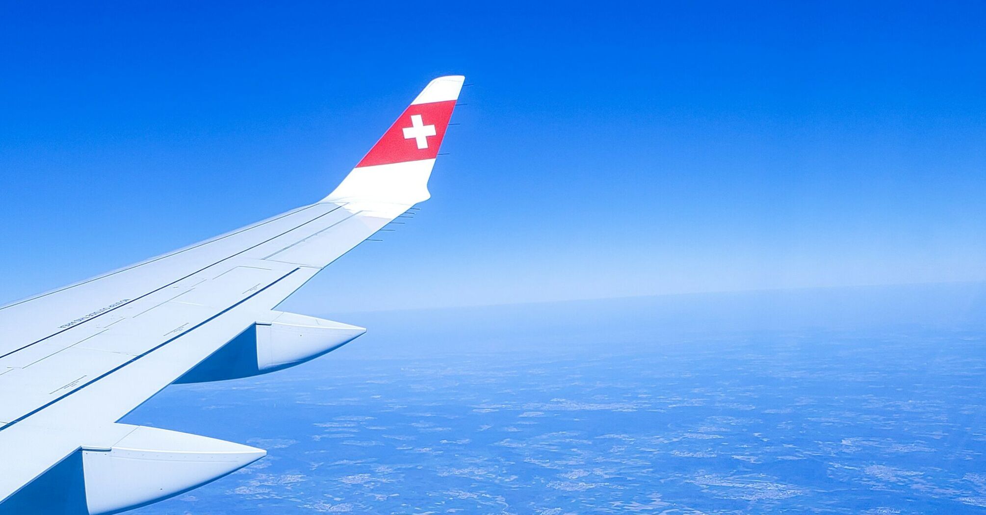 A SWISS Airlines wing in flight, soaring above the landscape with clear blue skies