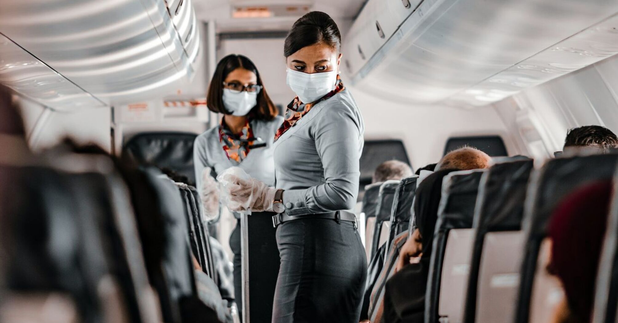 Flight attendants in a plane cabin wearing masks and gloves, serving passengers