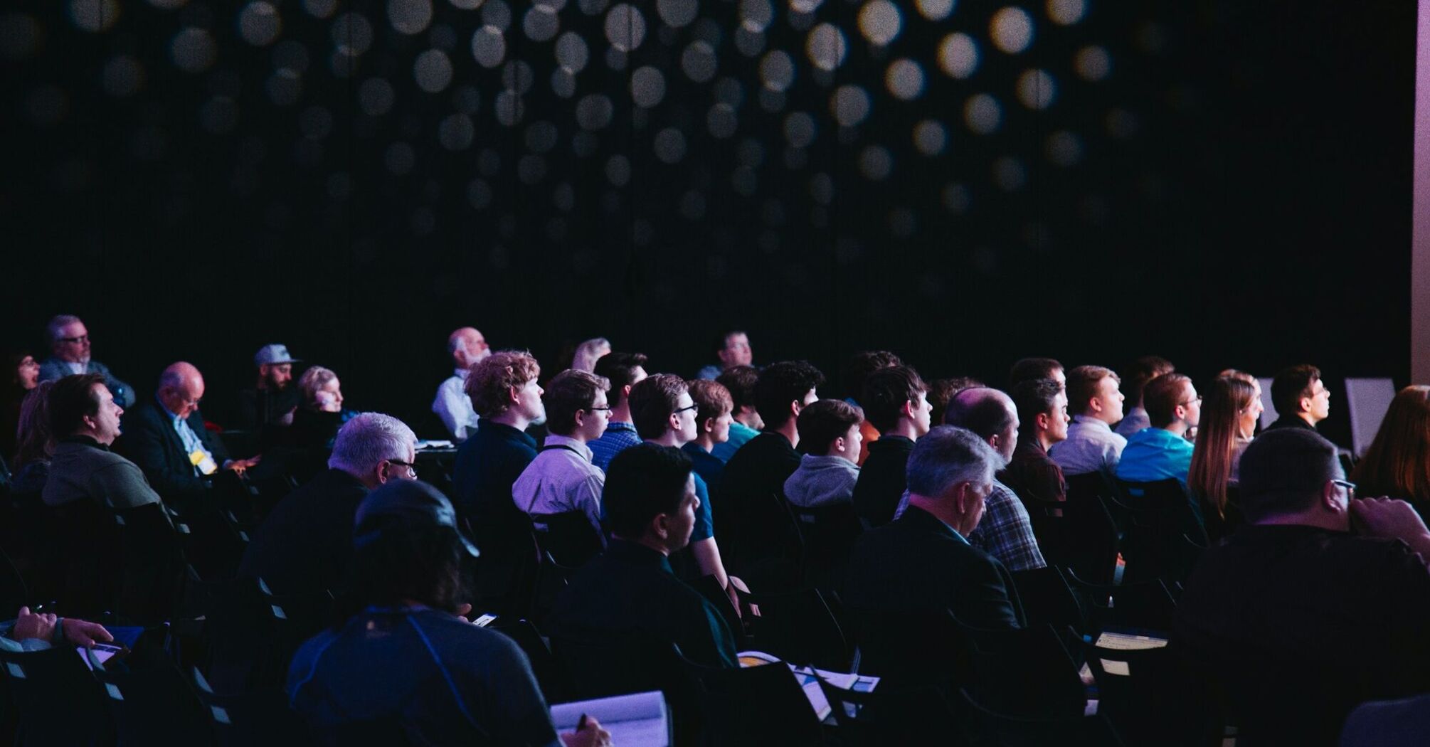 A diverse group of people attending a conference in a darkened room, focused on a presentation