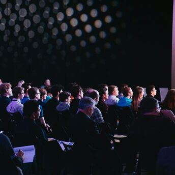 A diverse group of people attending a conference in a darkened room, focused on a presentation