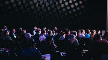 A diverse group of people attending a conference in a darkened room, focused on a presentation