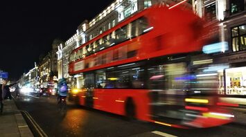 Blurred image of a red double-decker bus speeding through a city street at night, surrounded by illuminated buildings and cars