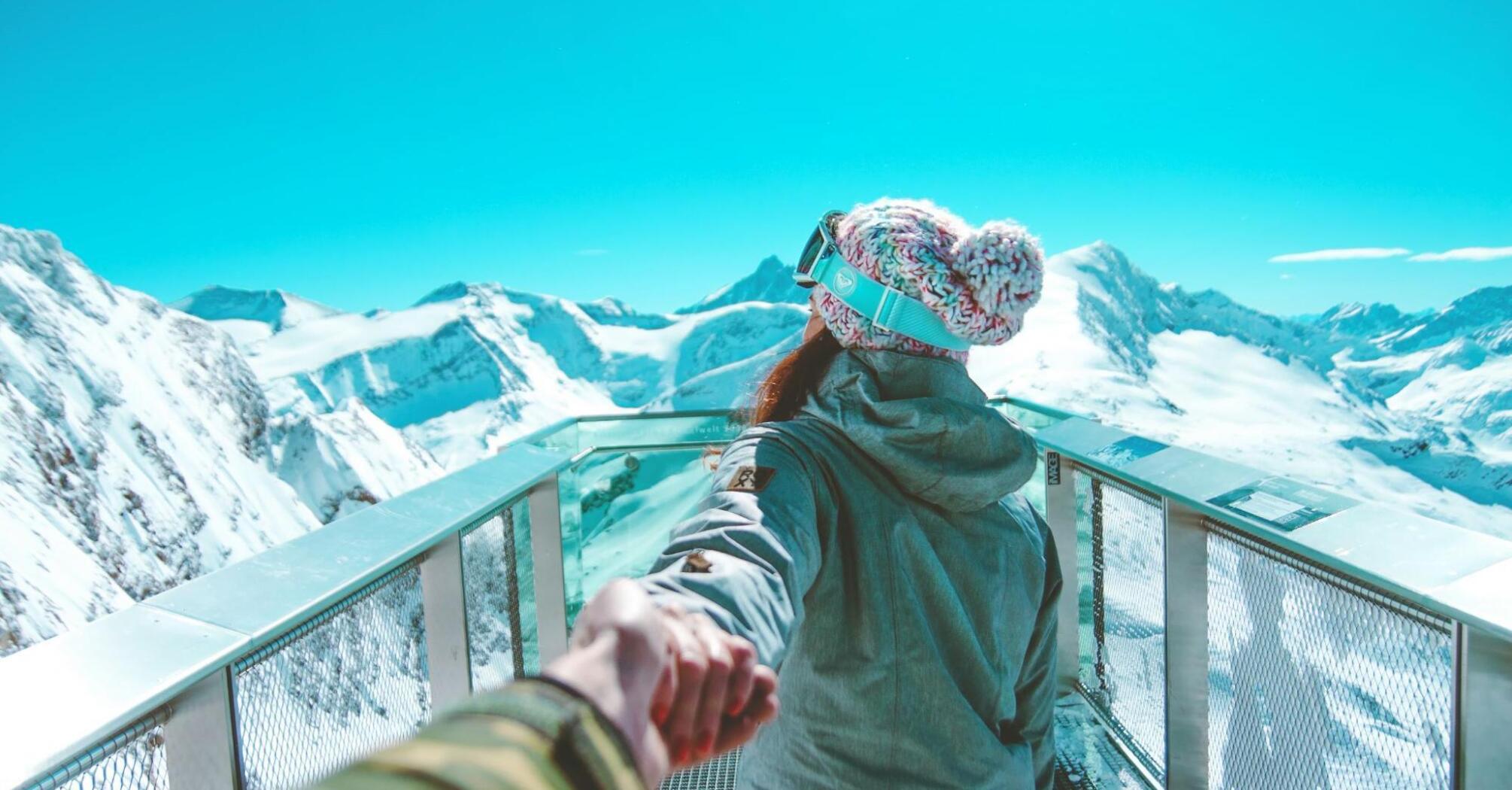 A person in winter gear holds someone's hand while standing on a snowy mountain viewing platform, with a stunning backdrop of snow-covered peaks and a clear blue sky