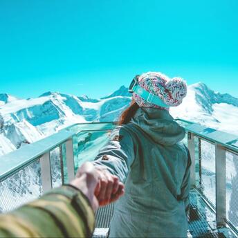A person in winter gear holds someone's hand while standing on a snowy mountain viewing platform, with a stunning backdrop of snow-covered peaks and a clear blue sky