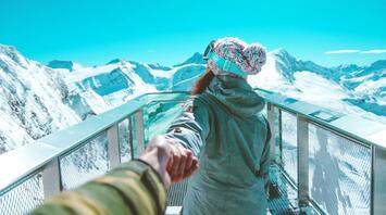 A person in winter gear holds someone's hand while standing on a snowy mountain viewing platform, with a stunning backdrop of snow-covered peaks and a clear blue sky