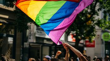 A colorful Pride flag waving in the air during a parade, held by participants in a lively, sunlit urban setting
