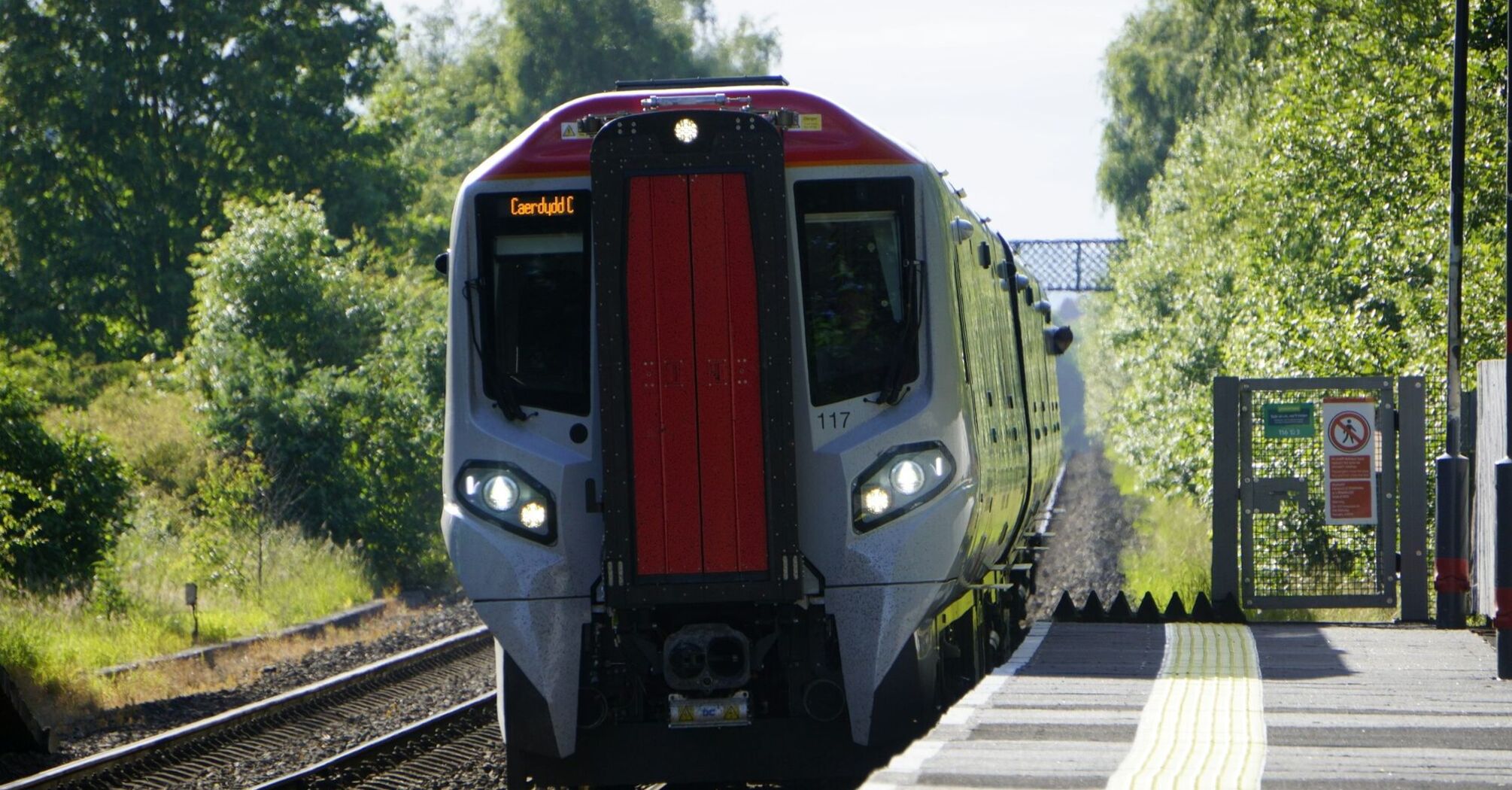 A modern Transport for Wales train arriving at a rural station under clear skies, with greenery on both sides of the railway track