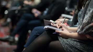 People taking notes during a conference or meeting, focusing on one person writing in a notebook