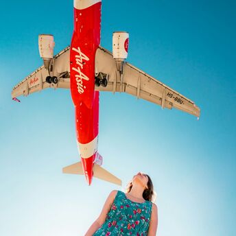 A woman in a floral dress looks up as an AirAsia airplane flies overhead against a clear blue sky