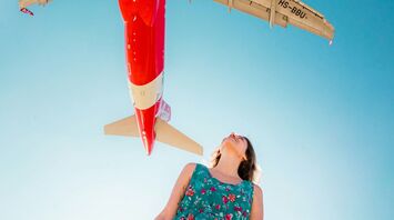 A woman in a floral dress looks up as an AirAsia airplane flies overhead against a clear blue sky