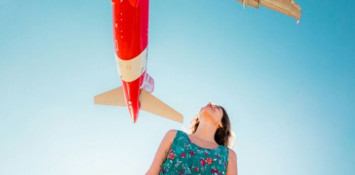 A woman in a floral dress looks up as an AirAsia airplane flies overhead against a clear blue sky