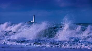 Waves crashing near a lighthouse under a dark blue sky