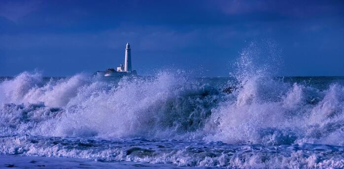 Waves crashing near a lighthouse under a dark blue sky