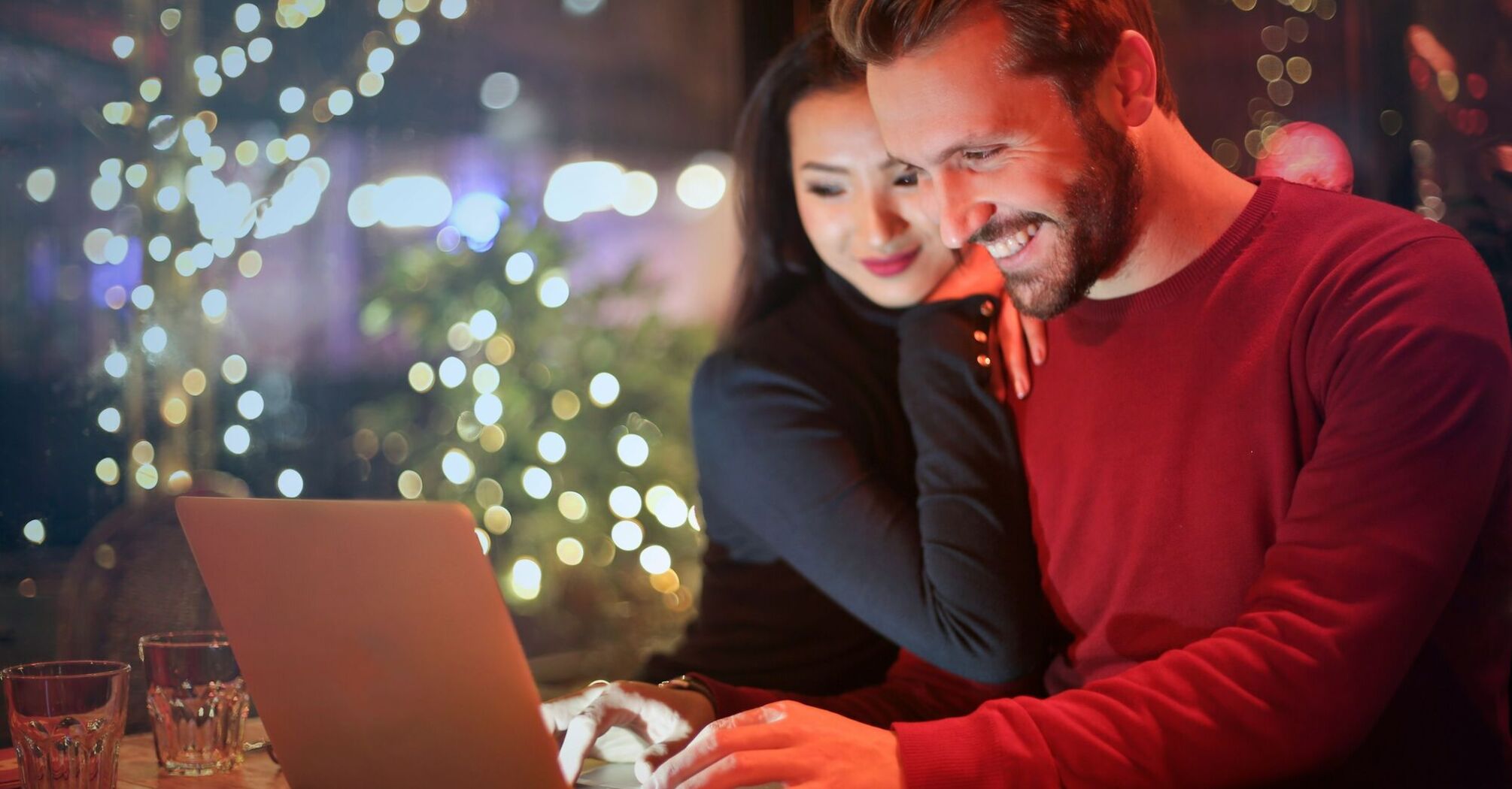 A smiling couple browsing on a laptop in a cozy cafe with festive lights in the background