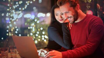 A smiling couple browsing on a laptop in a cozy cafe with festive lights in the background