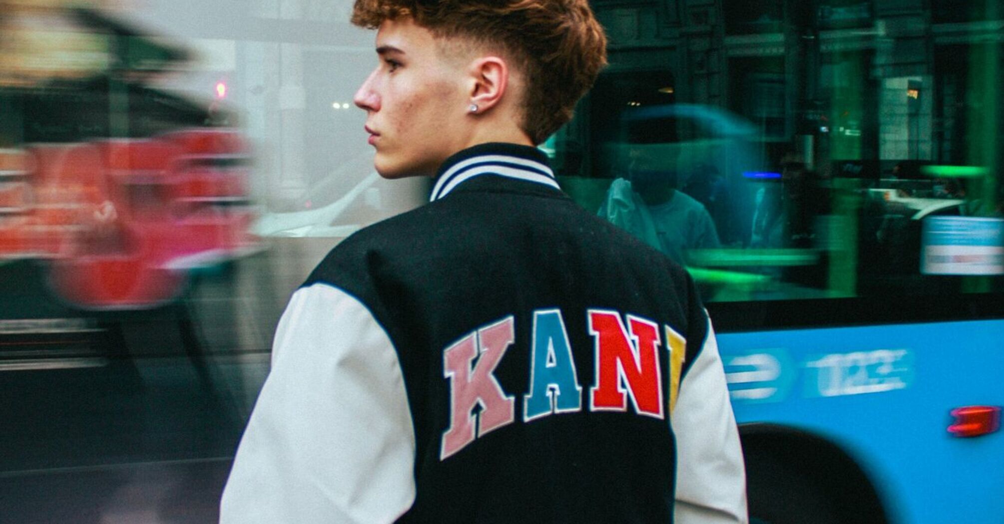 A young man standing on a city street in Madrid with a bus passing by in the background, showcasing urban transportation in Madrid